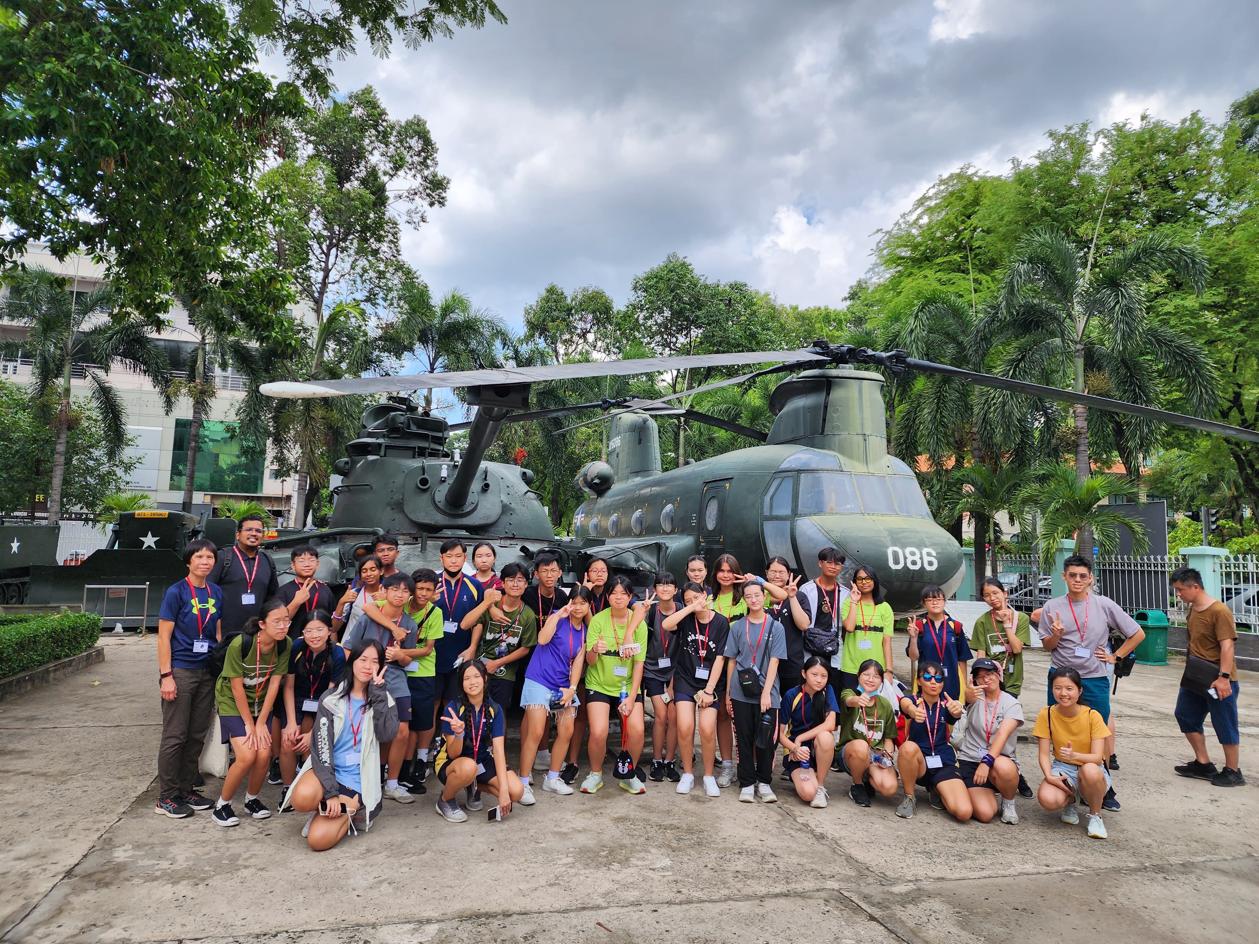 Group of students posing in front of a large green military helicopter, with trees and cloudy sky behind.